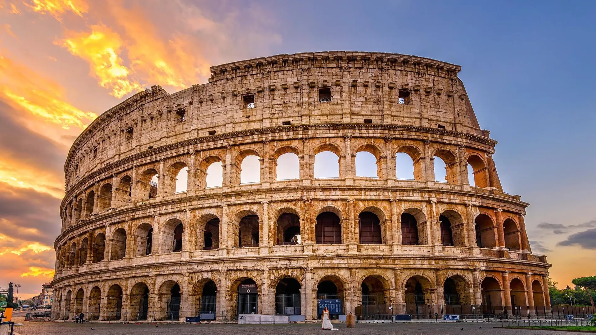 The iconic Gladiator in Rome with a beautiful sunset behind it and a woman in a white dress infront.