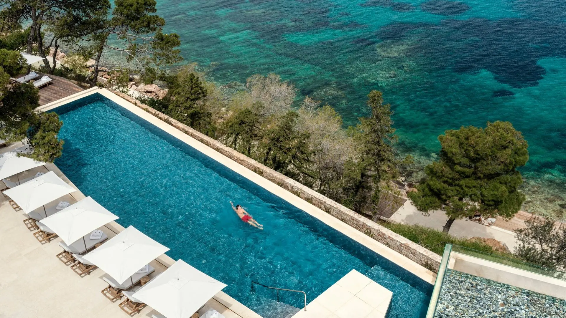 Bird perspective of a man in red swimming trunks swimming in a pool with white sunbeds and the ocean beside.